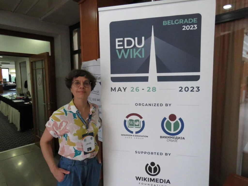 Heike Gleibs stands in front of a display stand bearing the inscription “EDU WIKI. Belgrade 2023” and the logos of Wikipedia & Education User Group, Wikimedia Serbia, and the Wikimedia Foundation.