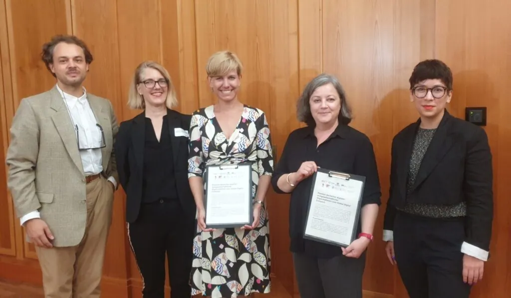 Group photo with five people. Two of them hold up documents.