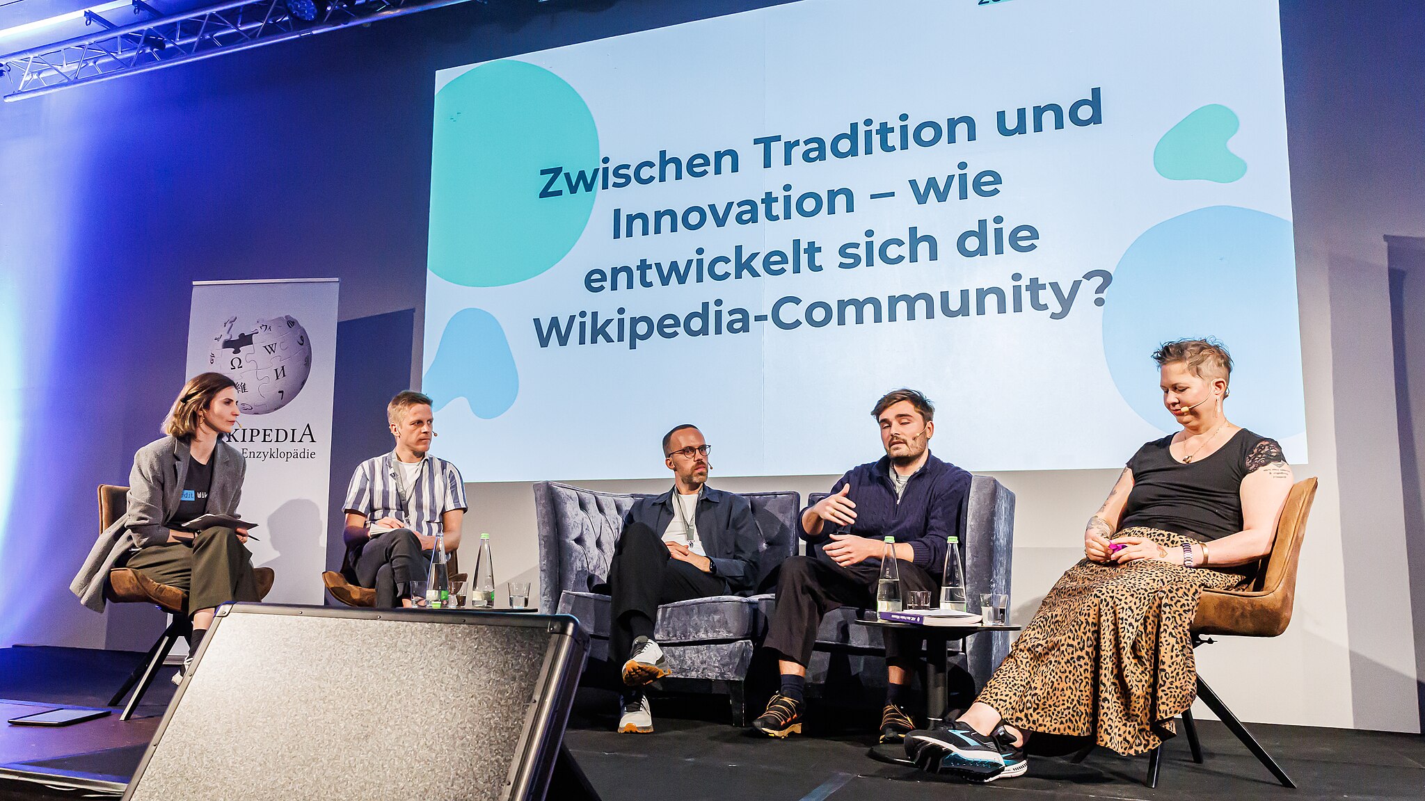 Fünf Menschen sitzen auf einem Podium. Hinter ihnen ist eine große Leinwand mit der Aufschrift „Zwischen Tradition und Innovation - wie entwickelt sich die Wikipedia-Community?“.