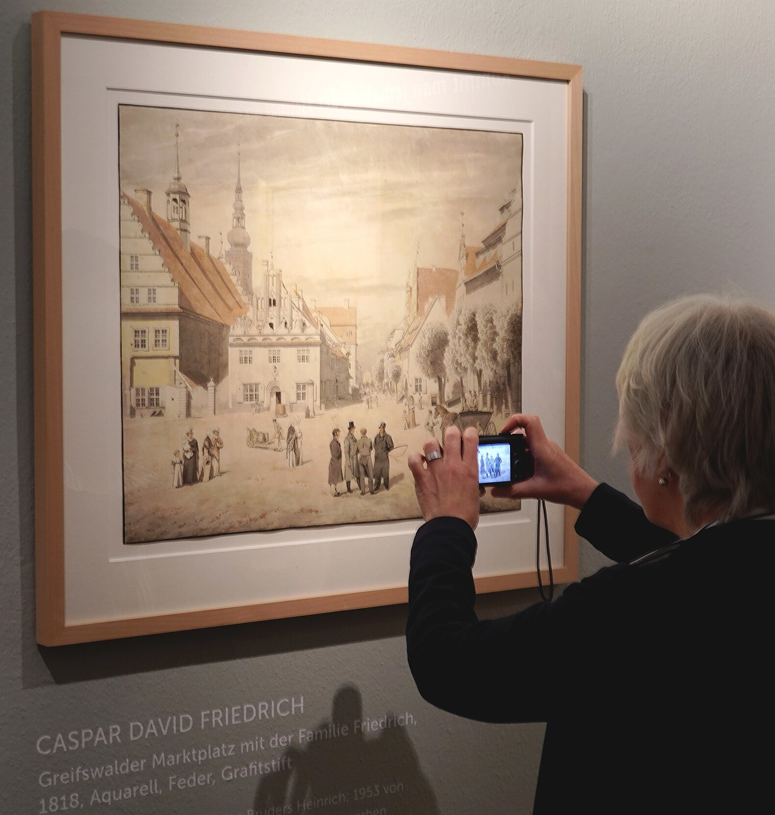 Eine Person macht ein Foto von Caspar David Friedrichs Gemälde „Greifswalder Marktplatz mit der Familie Friedrich“.