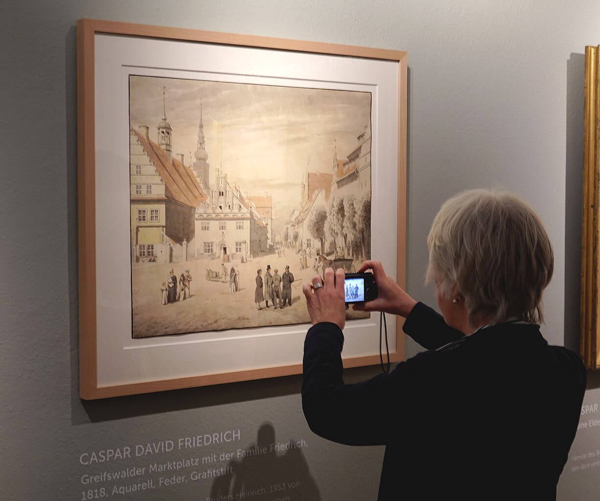 A person takes a photo of Caspar David Friedrich's painting ‘Greifswald market square with the Friedrich family’.