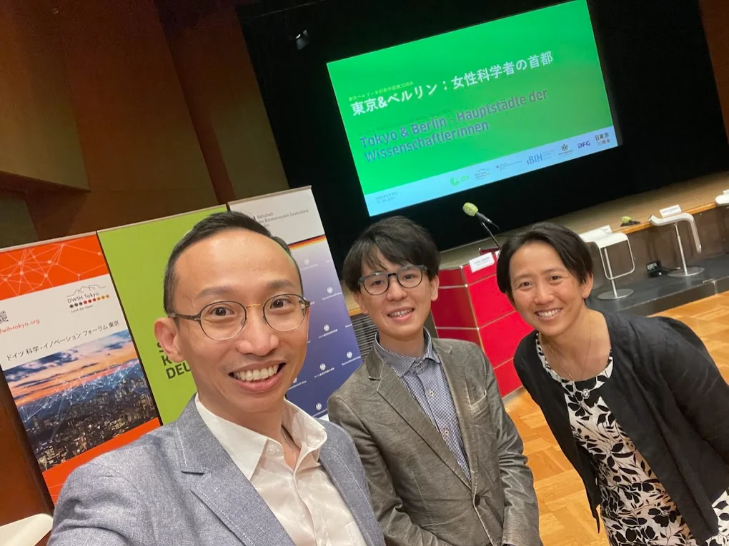 Ein Selfie von drei Personen vor einer Leinwand mit der Aufschrift „Tokyo & Berlin – Hauptstädte der Wissenschaftlerinnen“.