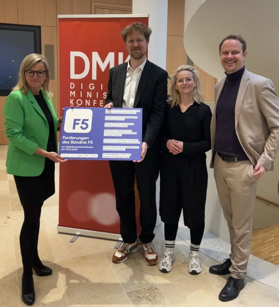 Vier Personen schauen in die Kamera und halten ein Plakat mit der Aufschrift „Forderungen des Bündnis F5“ hoch.