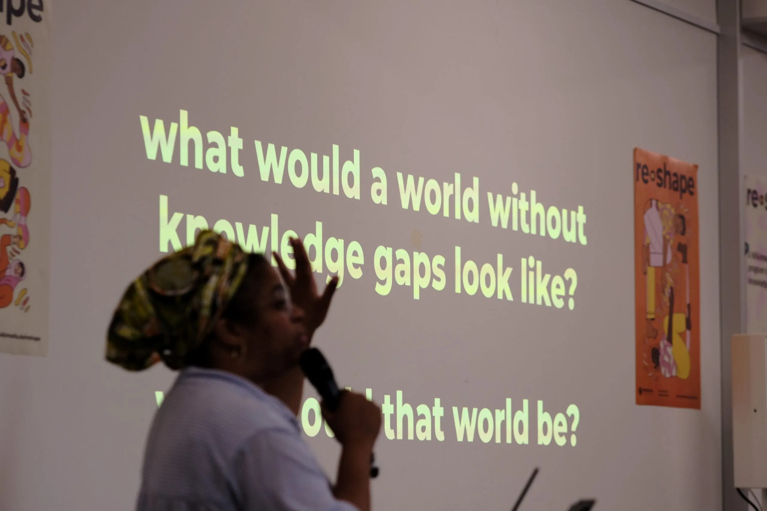 Eine Person spricht in ein Mikrofon und zeigt auf eine Projektion auf einer Wand. Dort steht: “what would a world without knowledge gaps look like?“