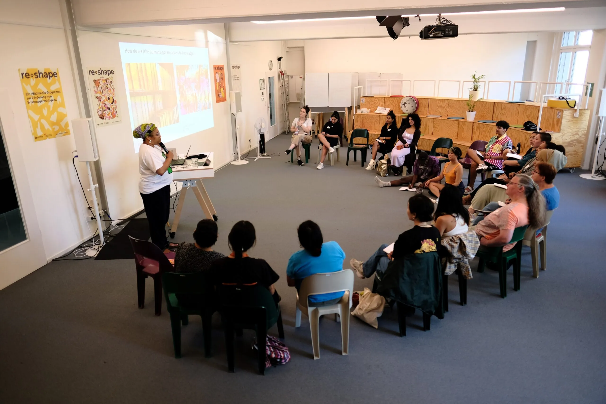 A semicircular circle of chairs. At the front, a person stands at a lectern and speaks into a microphone.