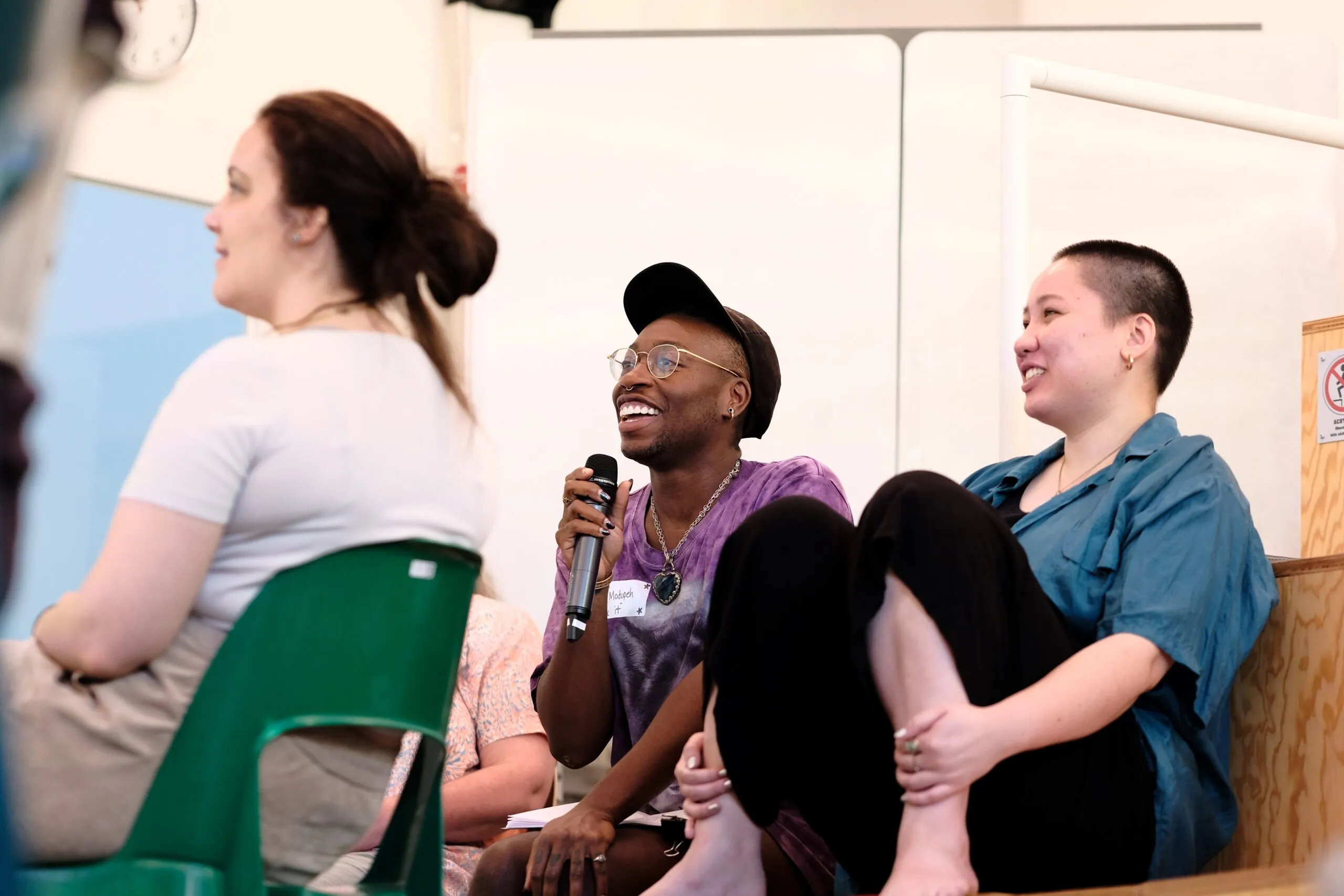 Two people sitting next to each other and smiling. The person on the left is holding a microphone.