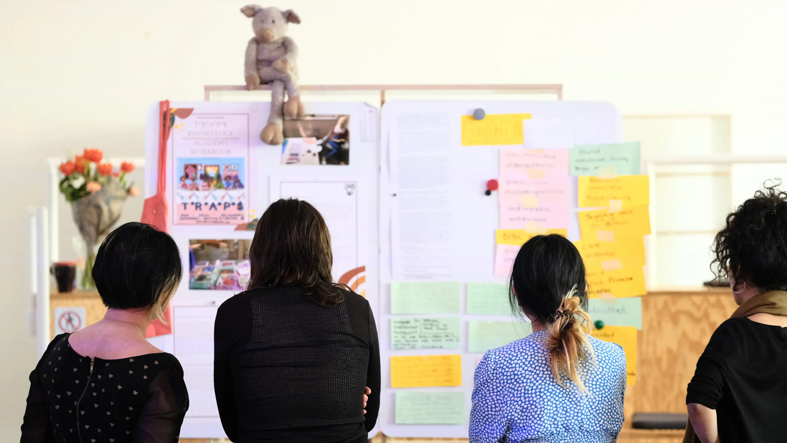 Vier Personen stehen mit dem Rücken zur Kamera und blicken auf ein Whiteboard mit bunten Zetteln.