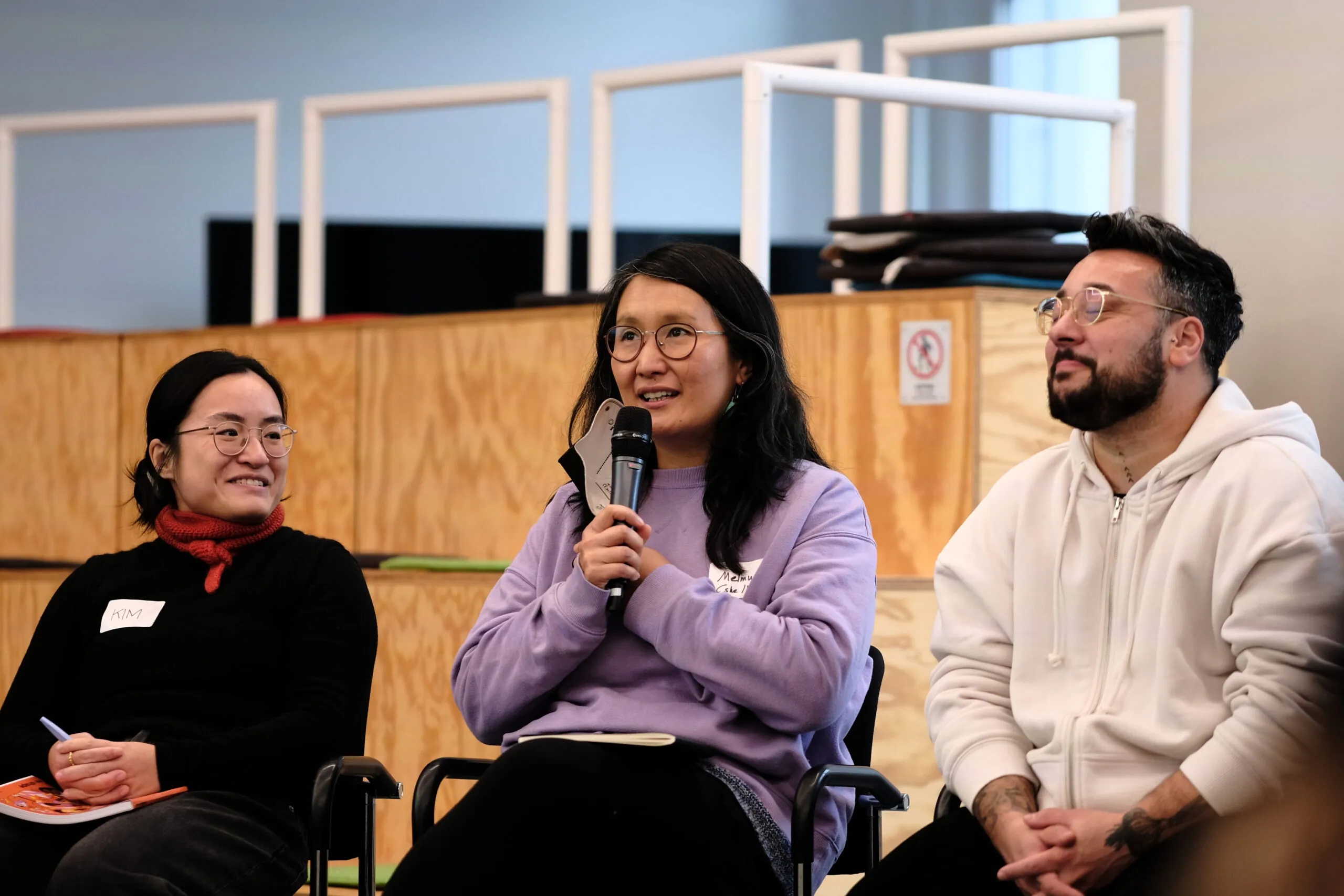 Three participants sit next to each other. The person in the middle holds a microphone in their hand.