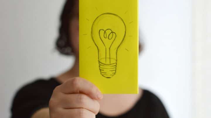 A person holding up a sticky note with a hand-drawn sketch of a light bulb on it representing thoughts, ideas and the sharing of lessons learned.
