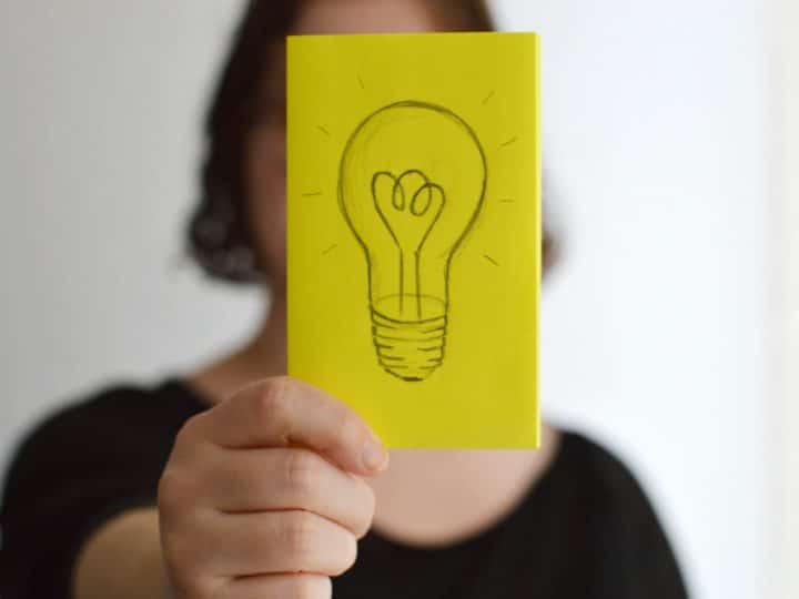 A person holding up a sticky note with a hand-drawn sketch of a light bulb on it representing thoughts, ideas and the sharing of lessons learned.