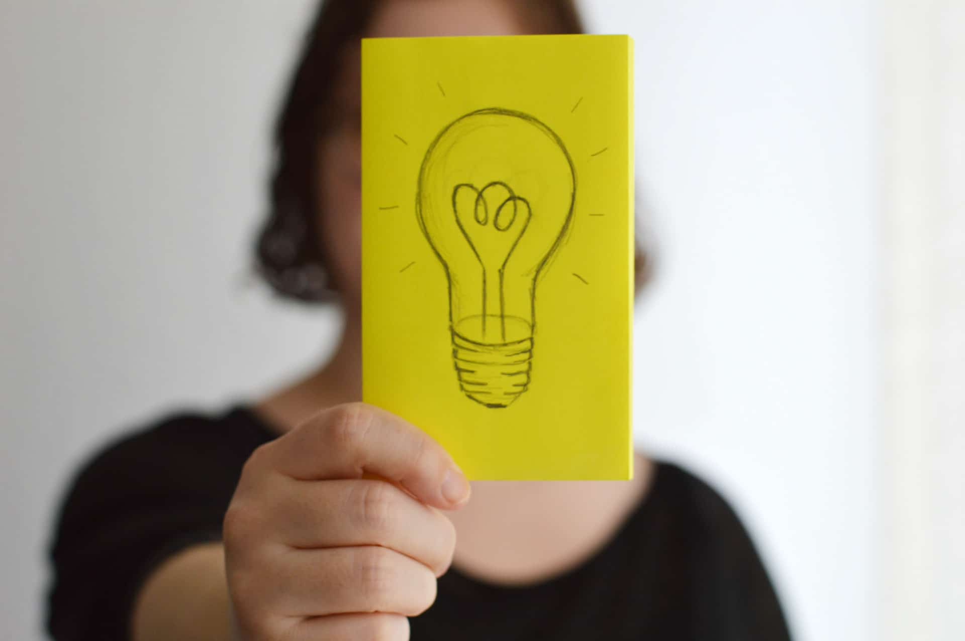 A person holding up a sticky note with a hand-drawn sketch of a light bulb on it representing thoughts, ideas and the sharing of lessons learned.