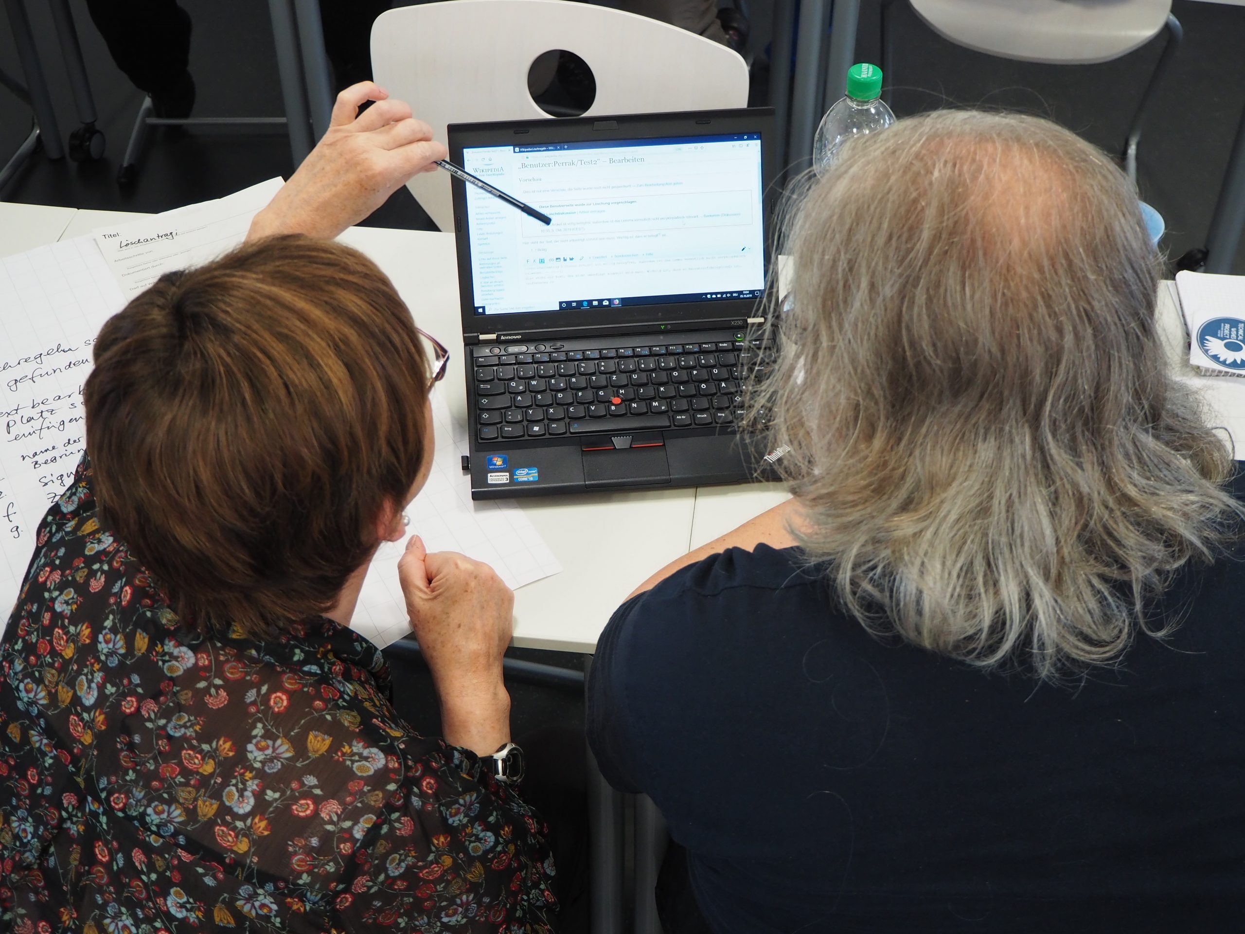 Zwei Personen sitzen nebeneinander am Tisch und schauen auf den Bildschrim eines Laptops und sind vom hinten zu sehen. Die Person links zeigt mit einem Stift auf etwas im Bildschirm.