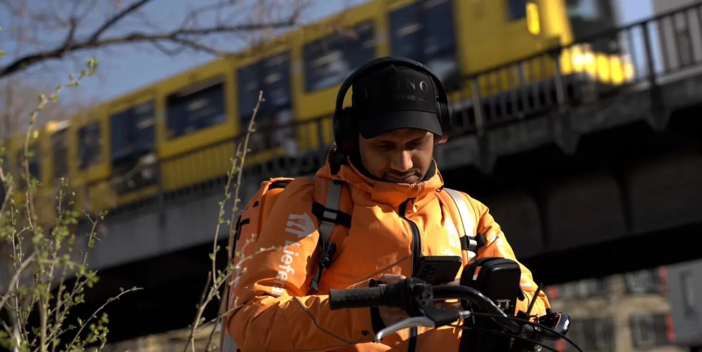 Ein Mann mit Lieferando-"Uniform" sitzt auf seinem Fahrrad und schaut auf sein Smartphone. Im Hintergrund fährt die Berliner U-Bahn übberirdisch.