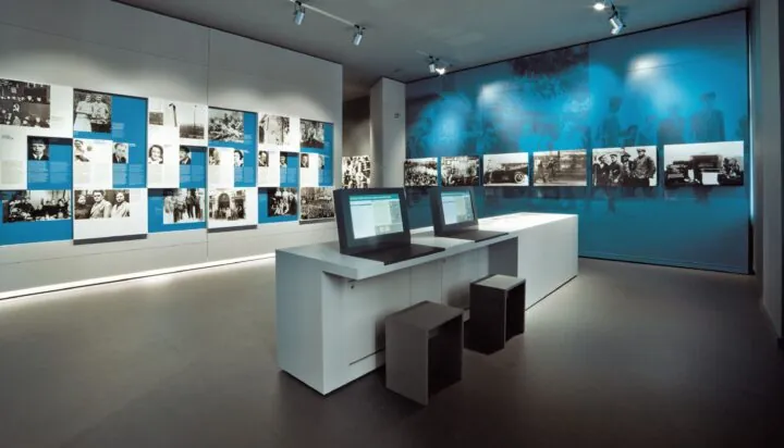 Eine Aufnahme der Daueraustellung. An den Wänden hängen schwarz-weiß Fotos und in der Mitte des Raums steht ein Tisch mit zwei Bildschirmen.