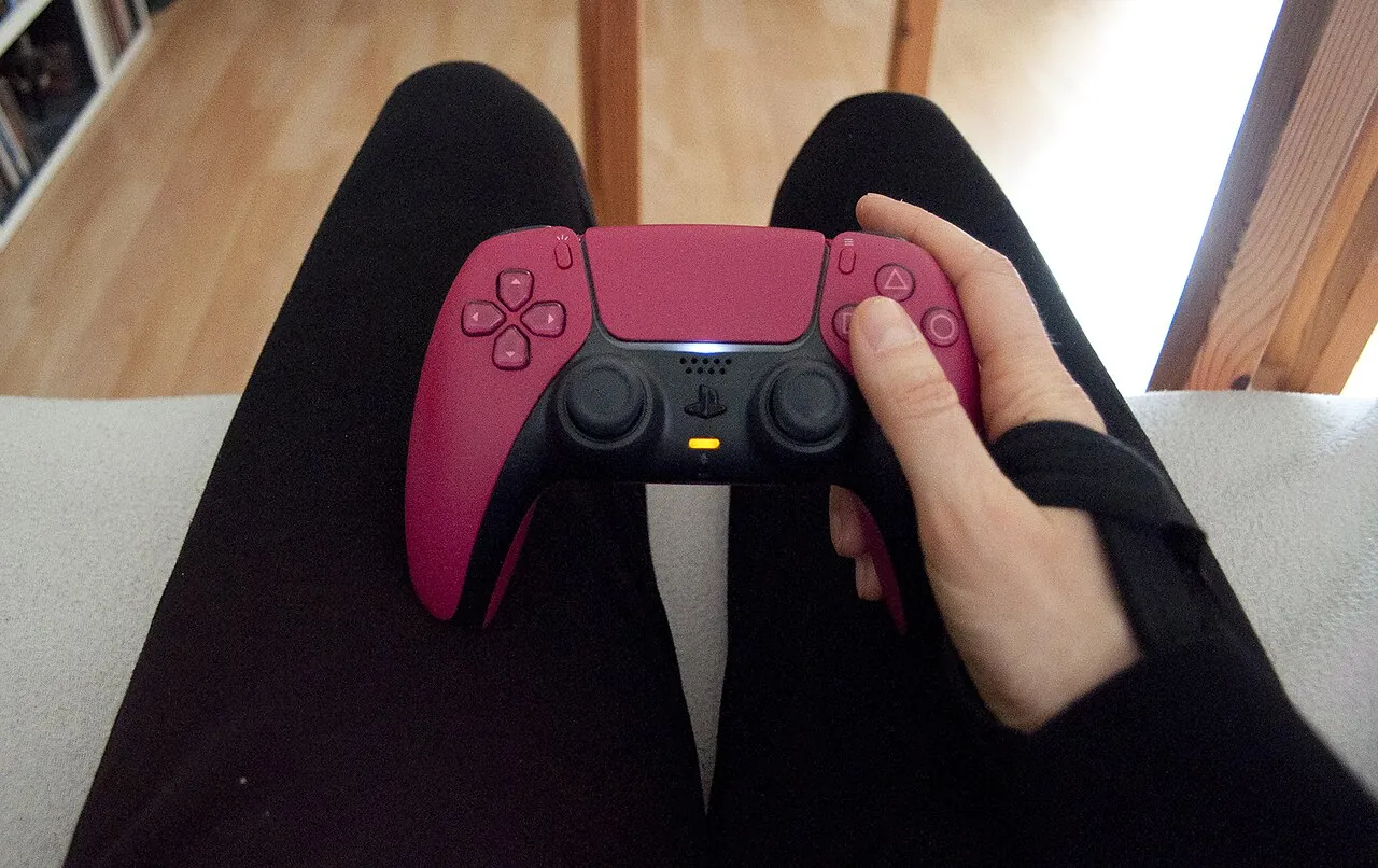 Eine sitzende Person hält in der rechten Hand mit Handgelenkbandage einen Playstation 5 Controller.